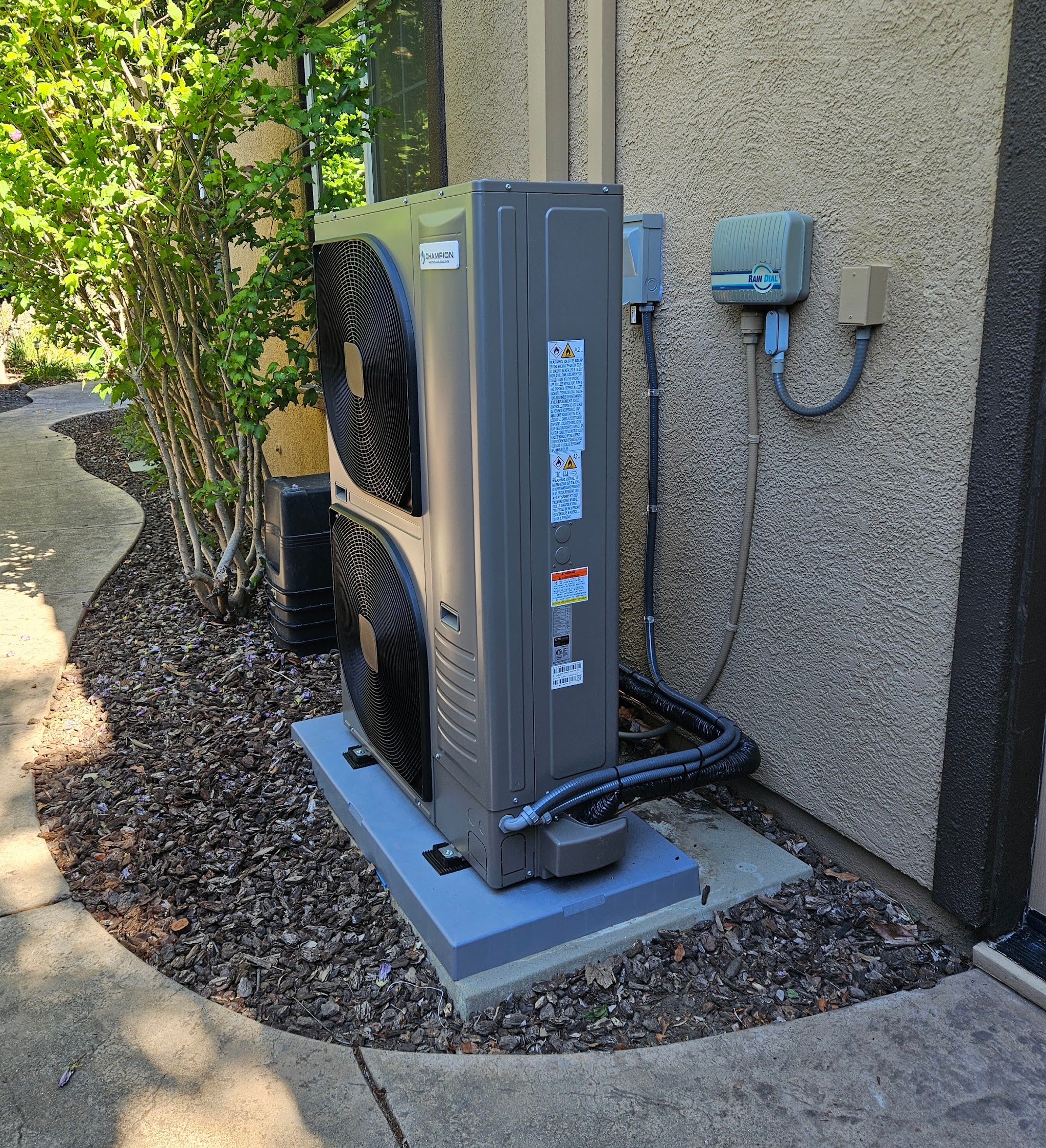 A newly installed dual fan heat pump condenser on a fresh concrete pad in a side yard, with tidy conduit and refrigerant line routing